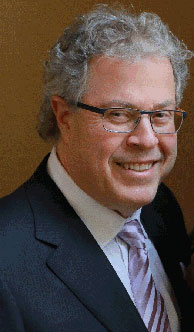 The image shows a man wearing glasses, with gray hair, dressed in a suit, smiling at the camera.