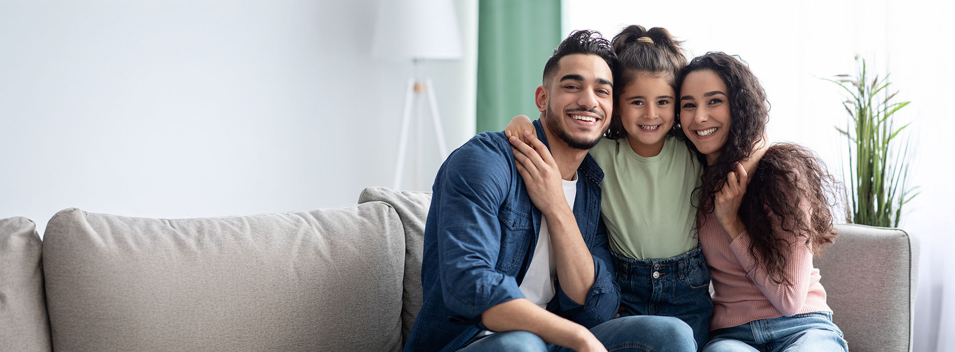 The image shows a family of three people - two adults and a child - posing together on a couch, smiling and looking at the camera.