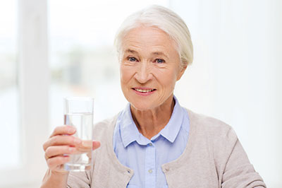 The image shows an elderly woman holding a glass of water with a smile on her face, suggesting a moment of health and well-being.