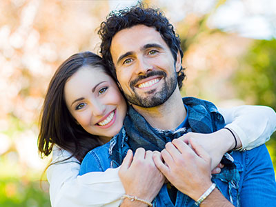 The image shows a man and woman embracing each other with smiles, set against a blurred outdoor background.