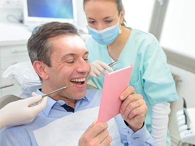 A man sitting in a dental chair while holding a pink card with a surprised expression, surrounded by dental professionals.