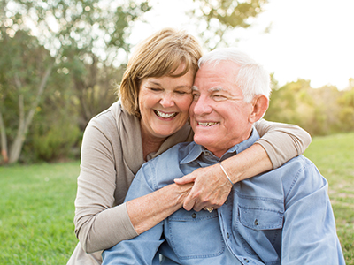 The image shows an older couple embracing each other outdoors during the day, with both individuals smiling at the camera.