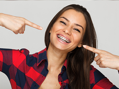 The image shows a woman with her finger pointing upwards, smiling at the camera, wearing a red plaid shirt and a blue jean jacket, against a white background.