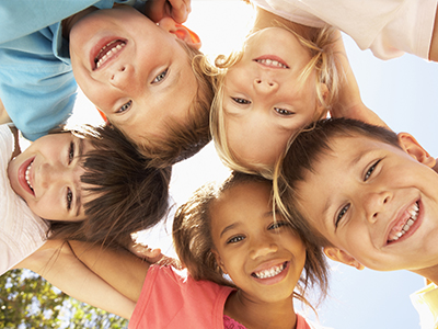 A group of children with different hair colors are smiling at the camera, posing together for a photo outdoors during the day.