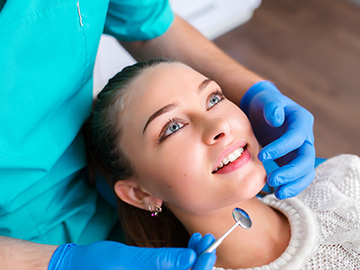 In the image, a young woman with blue eyes is seated in a dental chair while receiving a teeth cleaning treatment from a dentist.