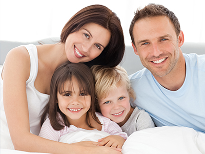 A family of four posing together on a bed with smiles, including a man, woman, and two children.