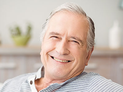 The image shows a smiling elderly man with gray hair, wearing glasses and a blue shirt, sitting comfortably with his hands on his lap in front of a window.