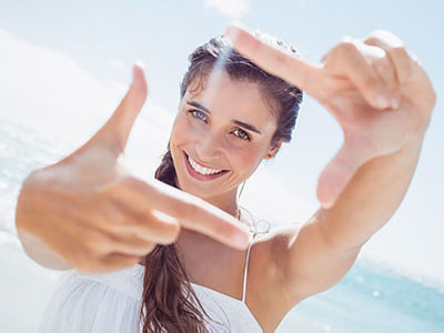 A woman with long hair smiles at the camera while holding up two fingers to frame her face, set against a bright background with clear skies and ocean waves.